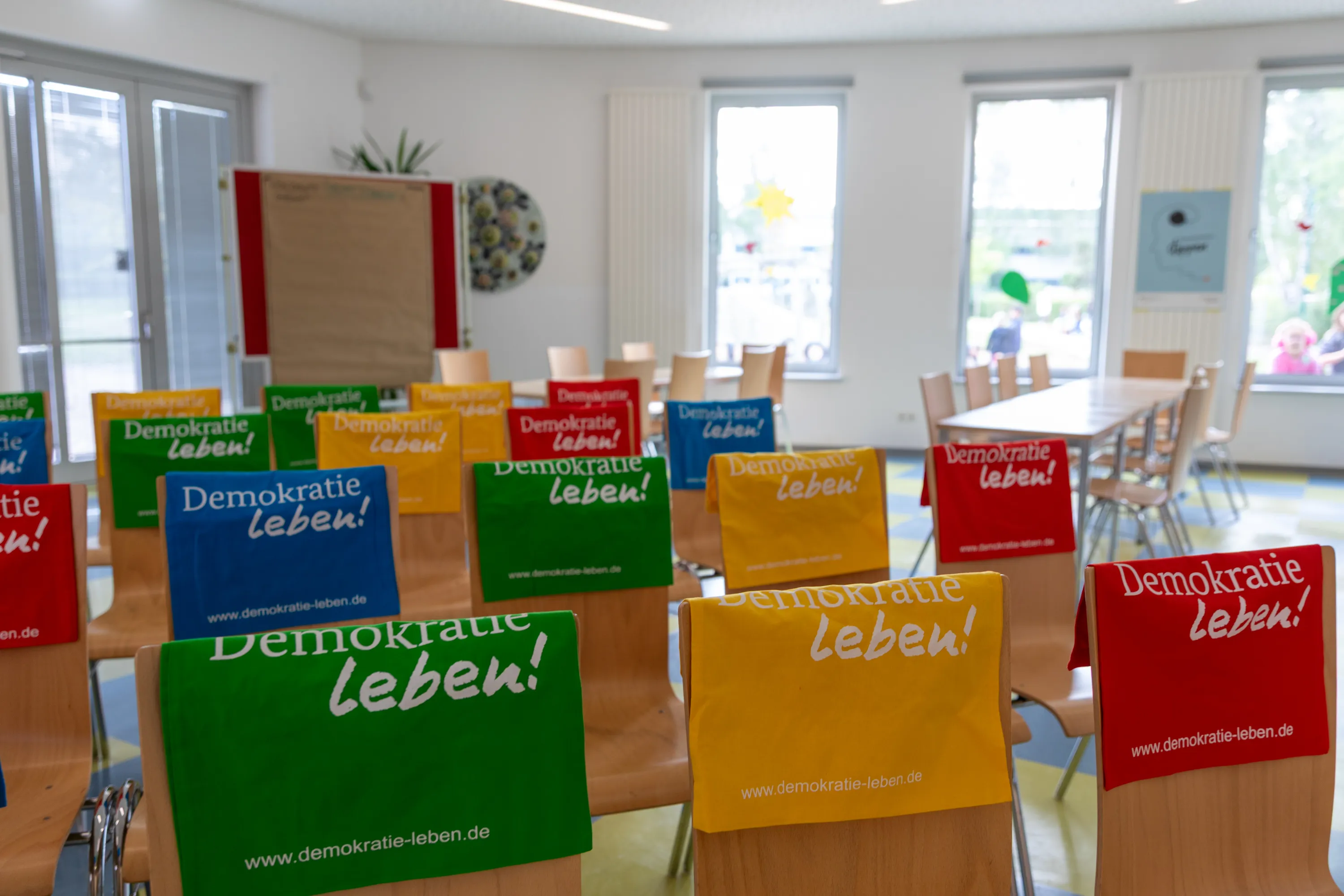 in einem hellen seminarraum stehen leere stuhlreihen mit bunten demokratie leben beuteln